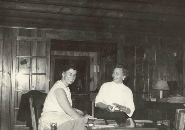 Marion Briggs (Mrs. Edward J.) Brumder, left, playing cards at a low table with Gladys Downey in a rustic cabin on Lake Julia. Knotty pine paneling covers the walls and there are exposed ceiling beams. Glass French doors open into another room. There are snowshoes on the wall to the right of the doorway.