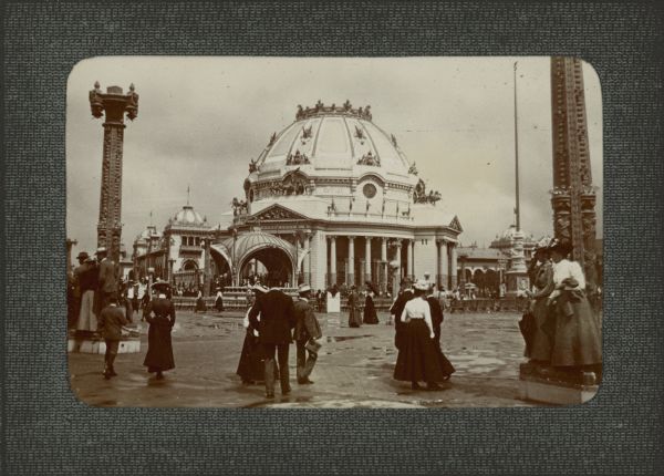 Well-dressed men and women are strolling the wet pavement at the Pan American Exposition in front of the Ethnology Building. Some people are standing on the bases of two light poles.