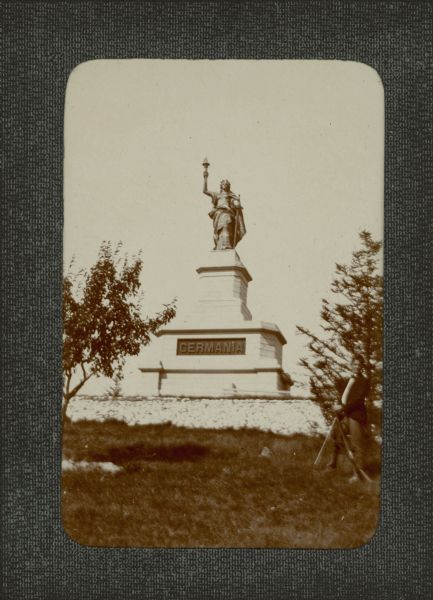 A young man, possibly a son of George Brumder, posing with a long gun near a statue of Germania. The sculpture is mounted on an elaborate stone plinth at Villa Henriette, the Brumder family summer home on Pine Lake.