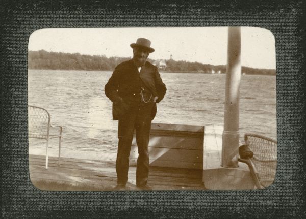 George Brumder, Milwaukee publisher and businessman, standing on his pier at Pine Lake. He is wearing a suit and hat. There is a low wall on the right, which supports a column. Two lightweight chairs are on the pier. A large house and tower are among trees on the far shorelinev. Below the photograph is written: "My Father Geo Brumder."