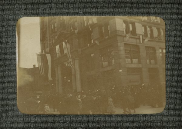 A crowd, including a newpaper boy on a bicycle, filling the sidewalk in front of the Germania Building on West Wells Street. There are numerous large and small American and German flags flying on the building in honor of the visit of Prince Henry of Prussia.  