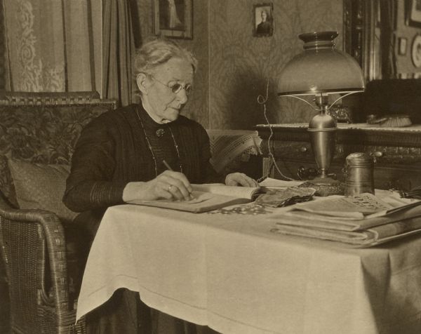 Henriette Brandhorst (Mrs. George) Brumder is sitting in an upholstered wicker chair at a small table writing in a book. There are coins and paper currency, newspapers, and a cylindrical metal container on the table. An electric light with a glass shade is sitting on the table. A handwritten captions states: "Grandmother keeping the books of the Frauenverein (Womens society)." A note on the reverse of the photograph further explains that Henriette was the head of the Frauenverein of Grace Lutheran Church.