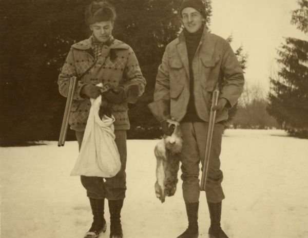 Art Frank, left, and Herbert Paul Brumder posing with game, carrying their shotguns "broken." Frank, who is smoking a cigar, is holding a bag from which he has pulled a ferret; Brumder is holding two rabbits. The ferret was likely used to track the rabbits. There is snow on the ground and both men are wearing heavy jackets, hats, and high laced boots. Each man is wearing one glove and one gauntlet. A written note on the reverse of the photograph identifies the setting: "at rear of George Brumder's Pine Lake home."