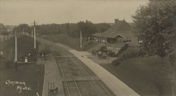 An elevated view of the Chicago, Milwaukee and St. Paul train depot at Nashotah. A woman is standing on the wooden sidewalk at left, near a small shelter. Horses, carriages, and a wagon are standing in front of the depot at right. There are utility poles along the tracks.  