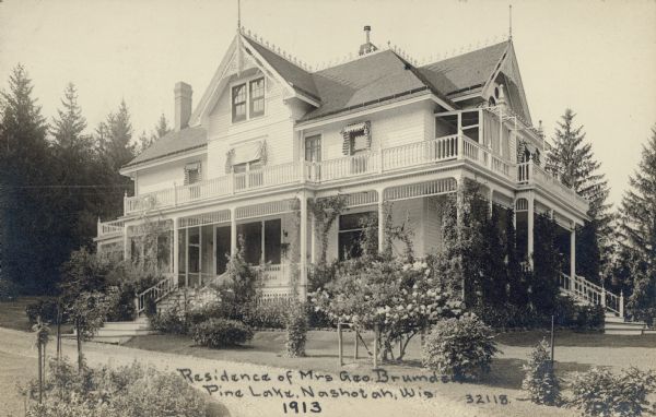 A photographic postcard of Henrietta Brumder's two and one-half story, wood frame summer home at Pine Lake. Henrietta was the widow of Milwaukee publisher and businessman George Brumder (1839-1910) who had the house built in 1893. Vines are growing on the large porches which are on three sides of the house, and there are awnings over the second floor windows. A large shrub is blooming in the yard and there are spruce trees in the background. The lake is out of view, to the right.  