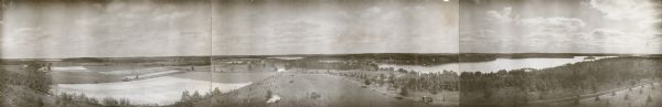 Three photographs taped together form a panoramic view from the top of the wooden tower built by George Brumder in 1892. The tower was built on Brumder's summer estate on Pine Lake. At far left (northwest) is Okauchee Lake, with Jacob Jacobson's house and barn standing to the left of two fields with corn shocks. The center photograph, facing east, shows, from left, North Lake, beyond Brumder's recently built barn; the smaller Mud Lake; and the northern end of Pine Lake. On the horizon beyond Mud Lake is the old white wooden church at Holy Hill.  Also shown at right in the center photograph is Brumder's Germania statue on its white stone plinth near the shore of Pine Lake, and in the foreground, a horse hitched to an empty carriage. The photograph at right, facing south, includes most of Pine Lake with its island and at extreme left, a glimpse of Beaver Lake. At extreme right is a similar glimpse of Nagawicka Lake. At left center of the right photograph is the roof of Brumder's summer home; his boathouse and gazebo are to the left of the main house.   