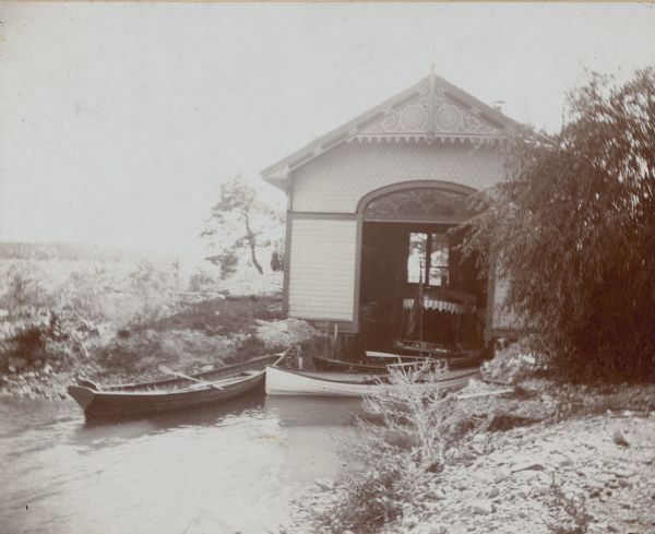 Three canoes, two of which have a square stern, are tied up outside of the boathouse at Villa Henrietta, George Brumdber's estate on Pine Lake. Inside the boathouse is a wood burning steam launch with canvas canopy. The boathouse has a decorative window above the door, and fretwork in the gable.  