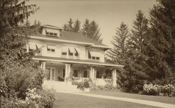 The east (lake-facing) side of the Pine Lake summer home of Henrietta Brumder, widow of Milwaukee publisher and businessman George Brumder (1839-1910). The two and one-half story home, originally built in 1893, was extensively remodeled. A porch extends along the entire length of the first floor. Awnings shade the upper windows. There are potted plants and vines on the porch; flowers are blooming in the yard.