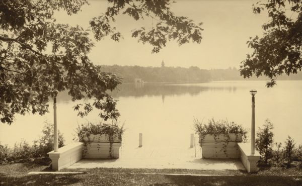 A view looking east over Pine Lake from the terrace in front of Villa Henrietta, the Brumder family summer home. In the foreground is a boat landing with low walls, flower boxes, and a light post on either side. A boathouse and tower are on the far shoreline.  