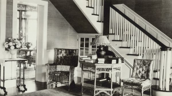 Two upholstered wicker chairs are flanking a matching table in the northwest corner of the living room of Henriette (Mrs. George) Brumder's summer home at Pine Lake. The open stairway frames the corner.  There is a vase of flowers on a side table next to a doorway on the left. Through the doorway is a view of the bright "dining sunroom."
