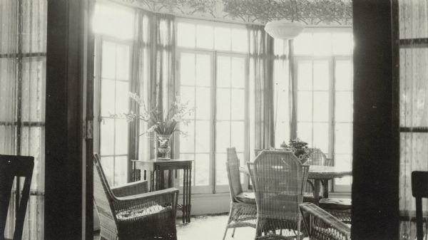 Light is pouring through tall windows into the "dining sunroom" of Henriette (Mrs. George) Brumder's summer home at Pine Lake. There is a trellis and foliage wallpaper frieze above the windows. The room is furnished with a wicker dining table and chairs and two wicker arm chairs. A side table is holding a vase of gladioli.