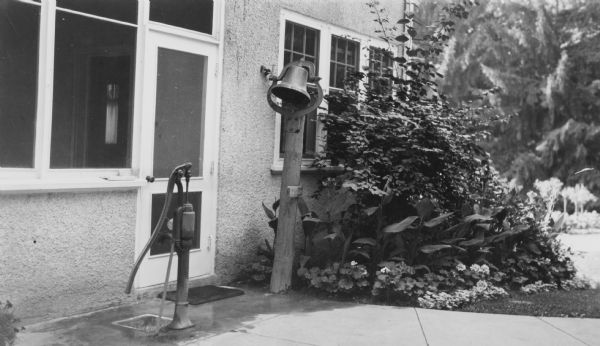 A hand pump and dinner bell flank the screen door of a porch at the Henriette (Mrs. George) Brumder summer home at Pine Lake. The walls of the house are stucco. Geraniums and cannas are growing at the base of a large shrub at right. A caption below the photograph reads: "The well pump and bell which was wrung [sic] loud and long for every meal."