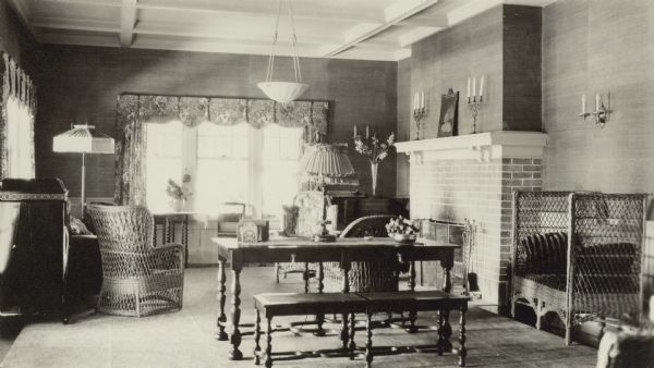An interior view, looking south, of the living room of Henriette (Mrs. George) Brumder's summer home at Pine Lake. An electric light is hanging from the coffered ceiling. The room is furnished with wicker chairs, a library table with a long bench, and a phonograph on the left. There is a fireplace on the right.