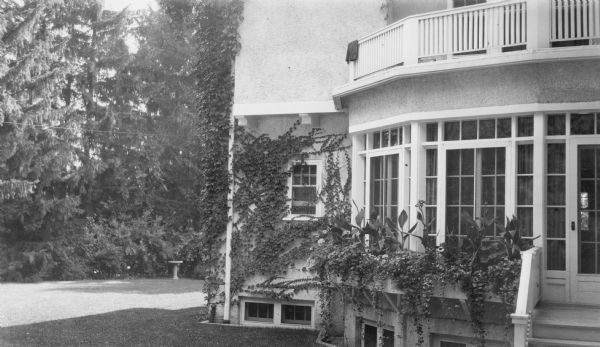 Geraniums and cannas grow in window boxes of the sunroom on the north side of Henriette (Mrs. George) Brumder's summer home at Pine Lake. There is a low railing around the balcony above. Vines are growing on the stuccoed walls of the house. There are large evergreen trees on the left and a birdbath on the lawn.
