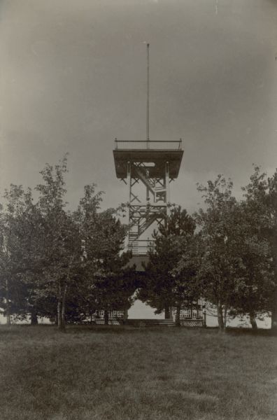 A wooden observation tower standing in a grove of trees. At the base is a stairway leading to a deck with a low railing. A tall flag pole rises above the deck at the top, and is supported by horizontal beams attached to the tower's uprights. A caption written below the photograph states: "On top of his hill George Brumder built a high tower in a cluster of pines affording a view of six lakes and miles of country."