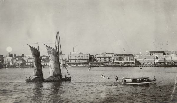 View across water towards a two-masted sailboat and a launch plying the waters of Havana Bay. Other boats are tied to the wharf in the background, with buildings behind.