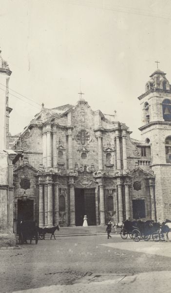 An unidentified woman is standing on the steps of the Havana Cathedral. She is wearing a hat, a long, light-colored dress, and dark gloves. There are several horse-drawn carriages on the plaza. Built of coral in the baroque style, the cathedral was completed in 1777.
