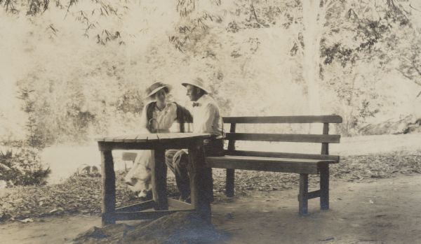 George F. and Thekla Brumder sharing a smile while sitting on a bench at a rustic table outdoors in Puerto Rico. Both are wearing light-colored clothing and hats.