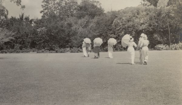 A group of men and women are walking across a broad lawn as they tour a garden in Puerto Rico. The women are carrying parasols. There is lush foliage in the background, including two palm trees.