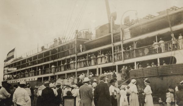 Passengers line the decks of the S.S. <i>Grosser Kurfürst</i>, docked at Colon, Panama, as others climb the gangplanks to board. A crowd is watching from the dock. Two lifeboats are on the right, and the flag of the German Empire is flying on the left.