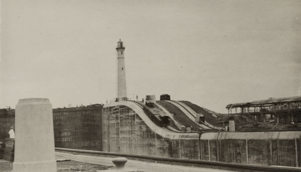 An unidentified man in light-colored uniform, on the far left just behind a column, is standing atop the near side of the Gatun Locks near Colon, Panama. On the opposite side, people are standing at the base of the newly constructed Gatun Northbound Rear Lighthouse. A "mule" trolley car is midway up the slope to the lighthouse.