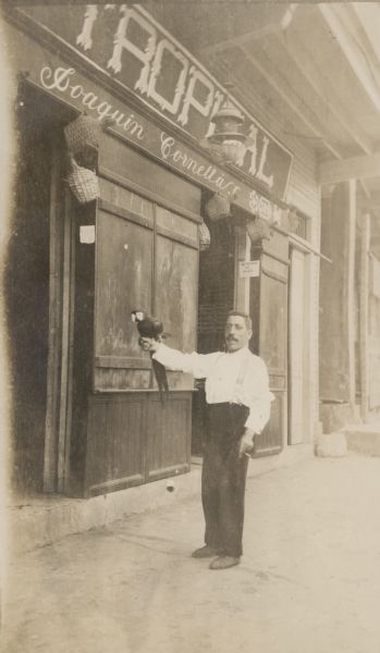 A man is standing in front of a store holding a macaw on his outstretched right arm. The man is holding a thin cigar in his left hand. A sign on the storefront reads: "Tropical" in large letters; below is written: "Joaquin Cornellas." The windows of the building are shuttered. Straw basket-covered bottles are hanging in the doorways.