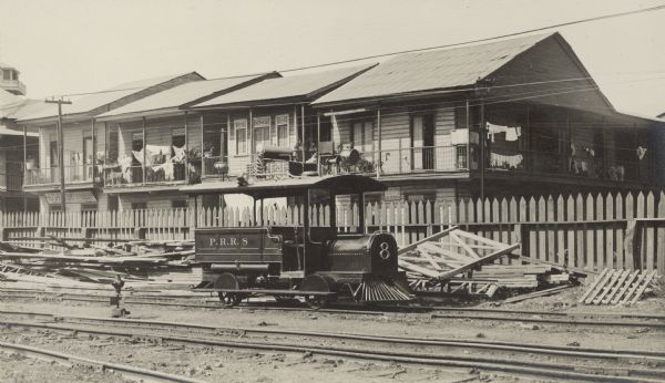 A small electric train engine identified as P.R.R. [Panama Railroad] 8 is parked in front of a row of two-story wooden houses. Lumber is piled near the tracks, which are separated from the houses by a picket fence. Laundry is drying on the second story porches. At left, at street level, is a restaurant identified on its sign as "La Martinequaise Cantine et Restaurant (Prix Modere)."