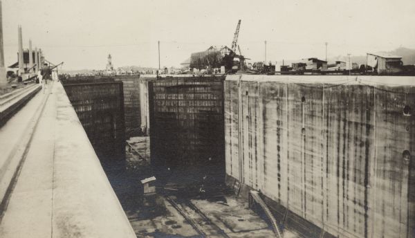 A view looking toward the Atlantic entrance to the Panama Canal, over the Gatun Locks during construction. The entrance lighthouse is seen in the distance. A man is standing at the edge on the left, and people standing on the lock gates in the distance are dwarfed by the massive walls of the locks. There is equipment on the floor of the locks and also atop the walls.