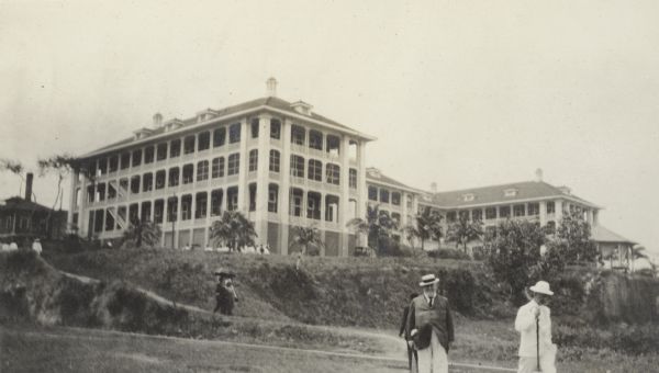 A stout, well-dressed gentleman is posing in the foreground, with the Tivoli Hotel behind him on a hill in Ancon, Panama. He is wearing a straw hat, jacket and tie. Another man in a light-colored suit and hat is walking near him on the right. The three-story hotel, built on a hill in 1906, has wrap-around porches on all levels and several vented cupolas. There are young palm trees in front of the hotel.
