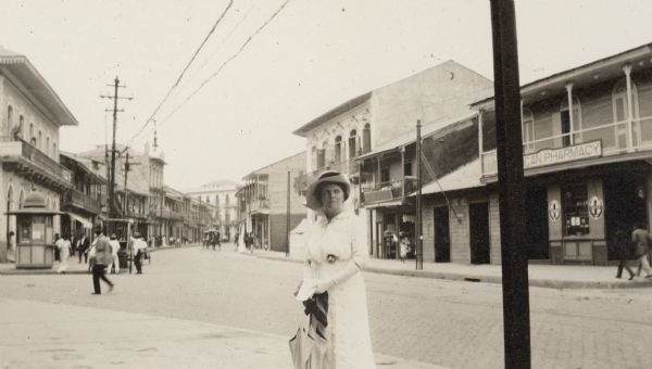 Thekla Wollaeger (Mrs. George F.) Brumder posing on a street in Panama, possibly in Panama City. She is wearing a long, light-colored dress, hat and gloves, and is carrying a parasol. Commercial buildings line the street behind her. There is a kiosk on the corner in the background on the left.