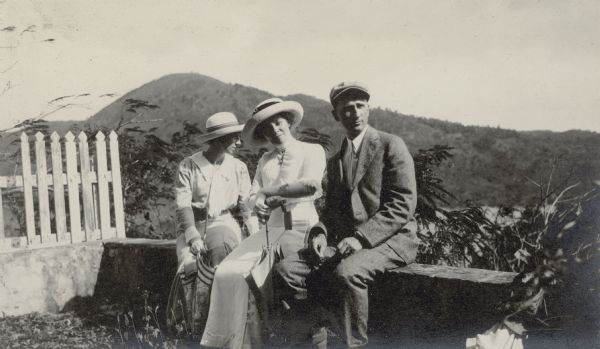 George F. Brumder and his wife, Thekla Wollaeger Brumder, center, are joined by an unidentified woman sitting on a stone wall. All of them are wearing hats, and the women, wearing gloves, are holding parasols. Behind them is a scenic view of St. Thomas, with a bay visible through the branches of a tree. There is a picket fence on the left.