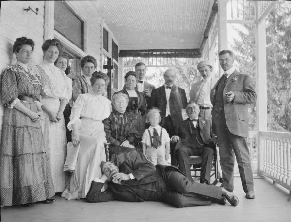 George Brumder, sitting at right, and his wife Henriette Brandhorst Brumder, sitting and wearing a dark dress, are posing with family members on the porch of their summer house on Pine Lake. Standing between George and Henriette is their youngest son, Herbert Paul Brumder. Another son, Herman Otto, is standing in the back row, wearing a light-colored suit and hat. Hugo Maercker (alternately spelled Merker), the husband of Ida Johanna Brumder, is reclining on his side in front of the group. At far right is George P. Mayer, husband of Amalie Brumder.