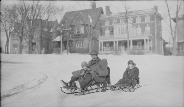 Herbert Brumder, pipe in mouth, is sitting on a sled with his son Philip George in front of him, and likely his son Herbert Edmund (face obscured) behind him. Daughter Barbara is sitting alone on a sled at right. Behind them are houses facing East Lafayette Place; a corner of their own house (2030 E. Lafayette Place) is glimpsed at far right.
