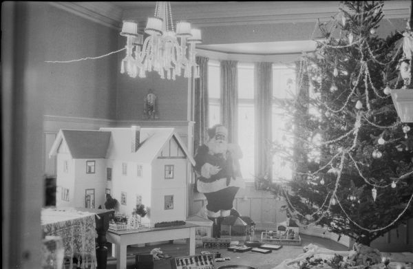 A tall Christmas tree, a cut-out Santa, and a dollhouse dominate a holiday display in front of the bay window in the dining room of Herbert Paul and Margaret Brumder, 2030 E. Lafayette Place. The tree is trimmed with tinsel garland and glass ornaments. A crystal chandelier with an extension cord running from one of the light sockets is hanging from the ceiling. Various toys clutter the floor.  