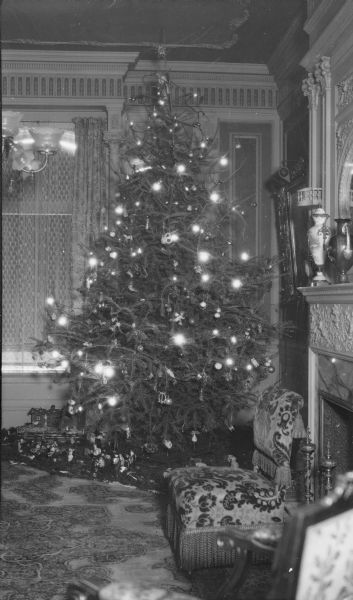 A tall Christmas tree with electric lights is standing in the ornately decorated parlor of the home of Henriette (Mrs. George) Brumder, at the corner of Grand (now Wisconsin) Avenue and 18th Street. A putz (nativity) scene with many characters is displayed under the tree. The tree is decorated with several styles of glass, paper, and wax ornaments. A portion of the fireplace and mantle is visible on the right. A chandelier fitted for both electric and gas lights is hanging from the ceiling.