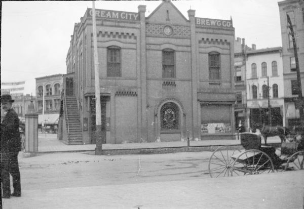 A view of the south (W. Wells Street) facade of Cream City Brewing Company's Triangle Bar. The two-story building features decorative brickwork. There is a double outside stairway on the left (N. 2nd Street) side of the building. Carriages and a horse are on the right; a bystander is looking at the camera from the far left. The building, demolished in 1919, occupied the triangle which is now Postman Square.
