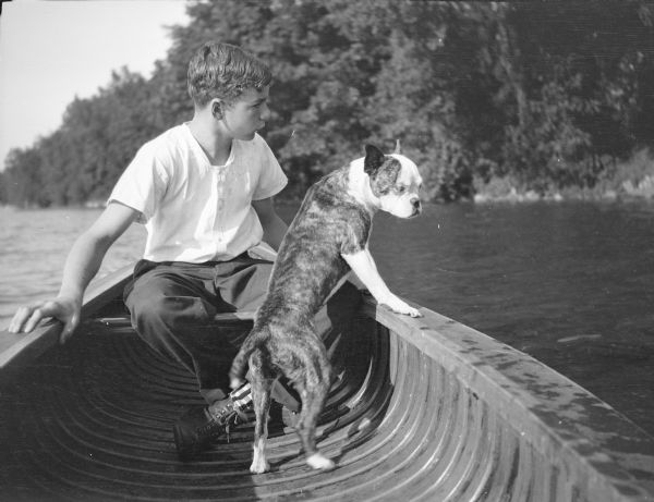 A boy, probably Edward John Brumder, sitting in a canoe with a Boston terrier-type dog. In the background is a shoreline.