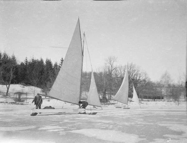 A man is bending forward near the mast of an iceboat as another man, wearing goggles, is standing to the left. There is a second iceboat on the right. A small amount of snow is partially covering the ice on Pine Lake.