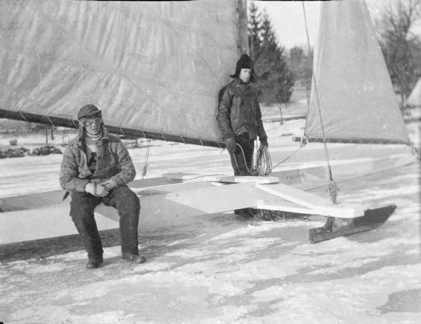 Two unidentified men posing with an iceboat on Pine Lake. The man sitting on the left is wearing goggles. "Reliance" is stenciled on the fore portion of the craft.