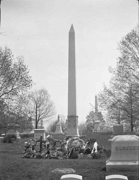 Floral tributes, including wreaths and a cross, form a large display at the base of the obelisk marking the Brumder family plot in Forest Home Cemetery.  Other monuments are visible, with names including Buckner and Pfister.  The trees are beginning to leaf out.  This image was created shortly after the burial of Milwaukee publisher and businessman George Brumder (May 24, 1839 - May 9, 1910).