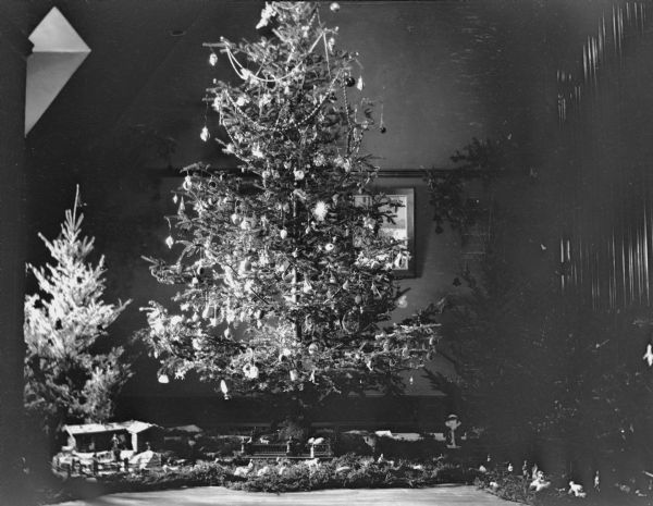 Light from a dormer window illuminates a number of Christmas trees, including a tall and lavishly decorated tree, in the home of George Brumder on Grand Avenue at 18th Street. The tree has an assortment of glass, paper, and tinsel ornaments, as well as strings of glass bead garland. A putz, or nativity scene, covers the floor among the trees, and includes a Santa Claus figure on the right.  A note on the negative envelope reads: "Christmas tree, Father's last Christmas, 1909." Brumder died May 9, 1910.  