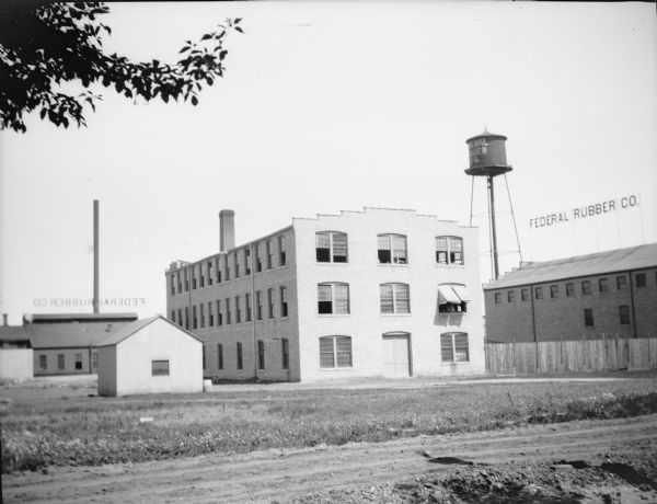 A water tower and two brick chimneys rise above the buildings of the Federal Rubber Company. These include a three-story brick building with large windows, center, and a smaller brick building at right. Large signs mounted on the roofs of two buildings identify the company. A small wooden shed is seen at left, and a larger building with clerestory windows is in the background. There is an unpaved drive in the foreground.