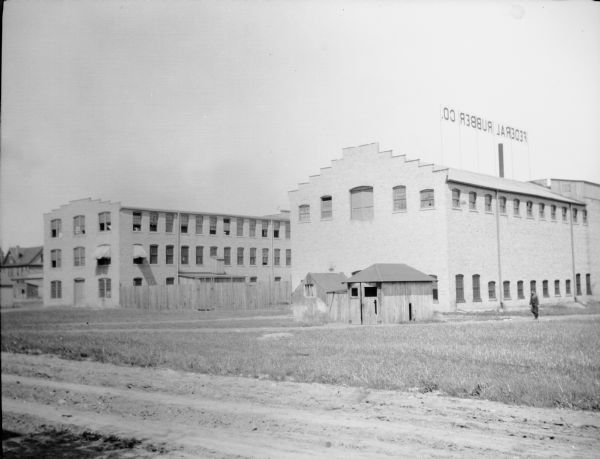 A large sign mounted on a brick factory building identifies the Federal Rubber Company. A second building, similar in size, is to the left, and there is a two-story house in the background. A man is walking on the right, in the direction of two small wooden sheds. There is an unpaved road in the foreground.