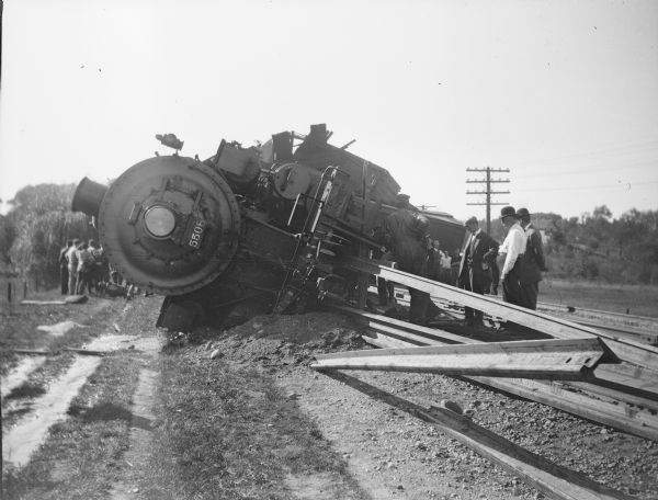 Milwaukee Road locomotive 5508 resting on its side after a derailment. The engine struck the rail, seen in center foreground, which had been driven upright into the ground to prevent trains from running onto an uncompleted portion of track. The engineer and fireman were able to crawl from the wreckage. Several well-dressed gentlemen at right are observing the scene.