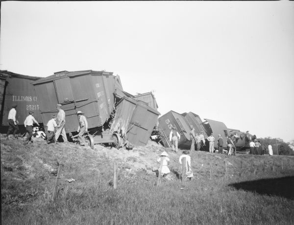 Railroad workers and a crowd of other men, women, and children standing and walking near the wreckage of a freight train. The engine and tender are lying on their sides, and at least three of the freight cars are derailed. There is a fence in the foreground.