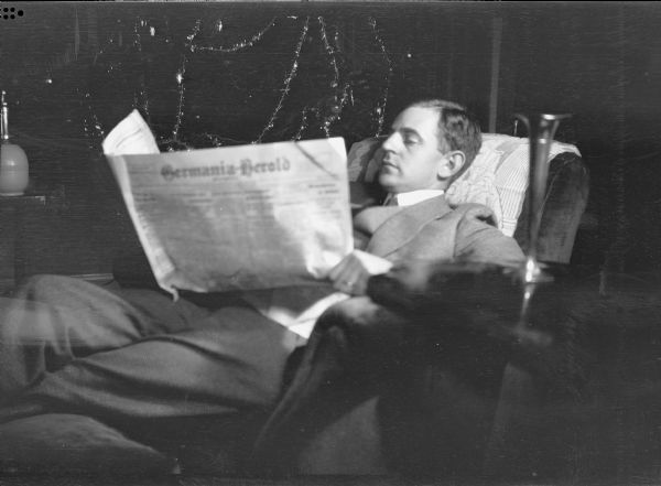 Herber Paul Brumder (1885-1967) relaxing in an easy chair with his crossed legs on an ottoman as he is reading the <i>Germania Herold</i> newspaper. There is a Christmas tree with tinsel garland in the background. A metal vase is sitting on a table in the right foreground. A note accompanying the negative reads: "Our first Christmas December 1916." Brumder and his wife Margaret Bouer Brumder (1893-1970) were married in July, 1916, and lived at 2030 E. Lafayette Place.