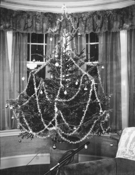 A Christmas tree decorated with glass ornaments, tinsel garland, and round electric lights is standing on a large block of wood in front of the bow window of the home of Herbert and Margaret Bouer Brumder, 2030 E. Lafayette Place. There is a vacuum cleaner under the tree and an easy chair at right.  