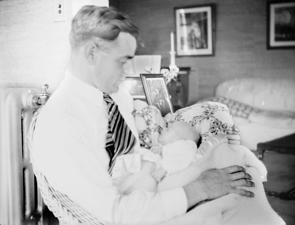 Herbert Paul Brumder, sitting in a wicker chair, is holding his infant son, Herbert Edmund Brumder. There is a steam radiator on the left.