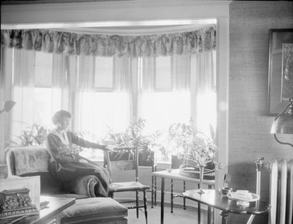 Margaret Bouer (Mrs. Herbert) Brumder (1893-1970) sitting on the arm of an easy chair near the bow window of her home at 2030 E. Lafayette Place. Her left hand is resting on a simple wooden chair. There are three fern stands with plants in the window, and a bouquet in a vase on a side table. A steam radiator is on the right.