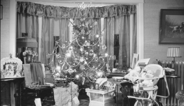 A night-time view of Herbert and Margaret Brumdber's living room decorated for Christmas. The live tree is decorated with electric lights, glass ornaments, tinsel ornaments and garland. There is a German paper pop-up Nativity scene at far left. Gifts around the tree include a Corona typewriter, wicker baby chair and folding high chair.